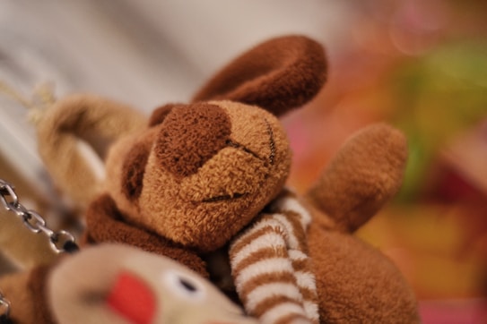 A soft, plush teddy bear with brown fur and striped limbs is pictured. It appears to be a stuffed toy in close-up, with blurred colorful elements in the background providing a warm and cozy setting.