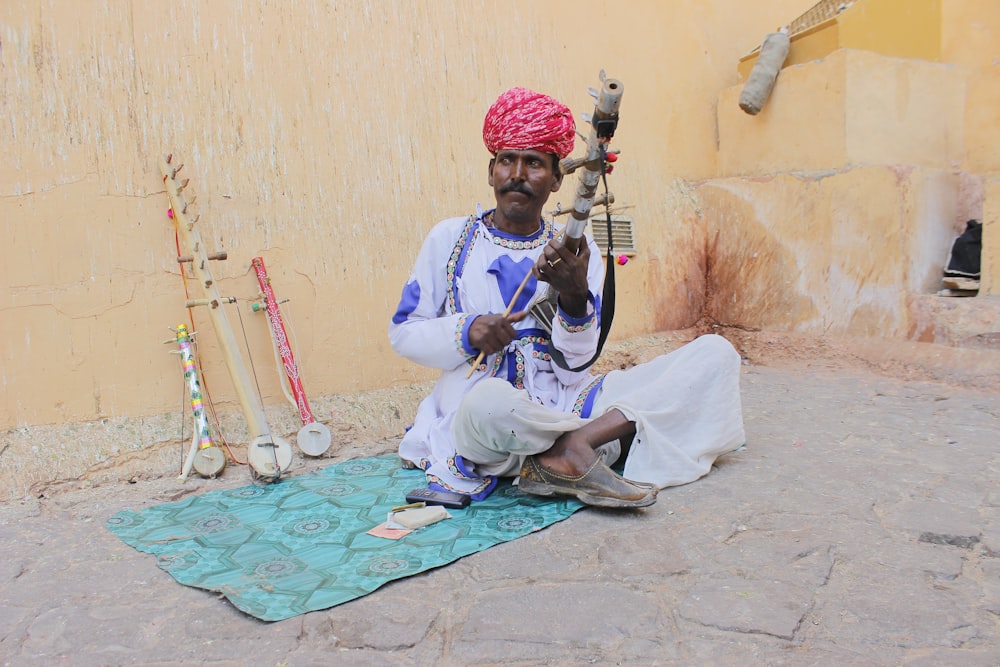 man in white thobe sitting on blue textile playing guitar