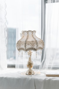 A vintage-style lamp with a detailed lace and fringe lampshade sits on a table by a window. The lampshade features intricate embroidery and bead details, while the lamp base has floral patterns. Soft natural light filters through sheer curtains, casting a gentle glow on the setup, creating a delicate and elegant atmosphere.