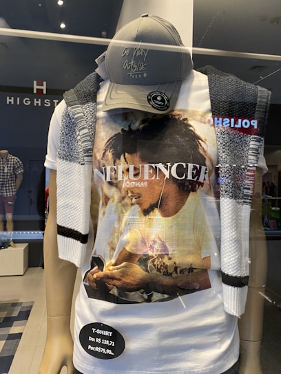 A store mannequin is dressed in a casual outfit consisting of a graphic t-shirt with a photo print of a person, a dark baseball cap with writing that says 'go play outside,' and a knit scarf draped over the shoulders. The store's name, HIGHST, is displayed behind the mannequin.