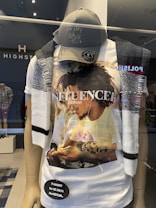 A store mannequin is dressed in a casual outfit consisting of a graphic t-shirt with a photo print of a person, a dark baseball cap with writing that says 'go play outside,' and a knit scarf draped over the shoulders. The store's name, HIGHST, is displayed behind the mannequin.
