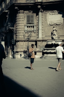 A city square with a historical stone building in the background, featuring architectural details like columns and sculptures. Two people walk in the foreground, one of whom is standing and wearing a hat, shorts, and a bag, while another person in white walks past.