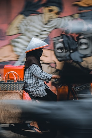 A person wearing a conical hat and a checkered shirt rides a motorcycle. The motorcycle is loaded with bags and packages. A colorful, abstract mural serves as the background, depicting artistic figures and shapes.