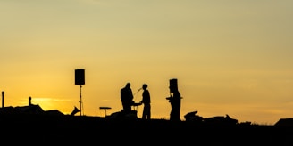 A silhouette of a band performing outdoors against a sunset sky with orange and yellow hues. The scene includes three people with musical instruments and microphones, as well as two loudspeakers. The figures are positioned on a slight incline, suggesting an open-air location.