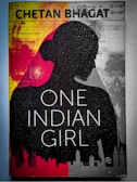A book cover featuring the silhouette of a woman's profile against a background divided into red and gray sections. The title 'One Indian Girl' is prominently displayed in large white letters, and the author's name is at the top in bold text. There are abstract graphics and faint images of cityscapes along the bottom edge.