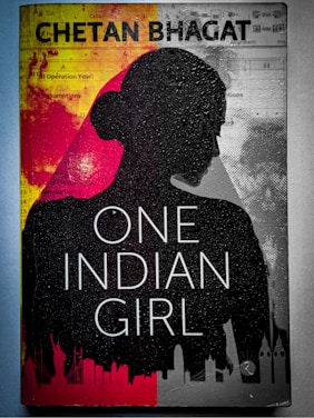 A book cover featuring the silhouette of a woman's profile against a background divided into red and gray sections. The title 'One Indian Girl' is prominently displayed in large white letters, and the author's name is at the top in bold text. There are abstract graphics and faint images of cityscapes along the bottom edge.