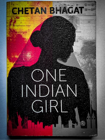 A book cover featuring the silhouette of a woman's profile against a background divided into red and gray sections. The title 'One Indian Girl' is prominently displayed in large white letters, and the author's name is at the top in bold text. There are abstract graphics and faint images of cityscapes along the bottom edge.