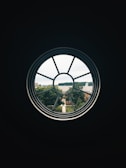 A circular window frames a view of a suburban neighborhood, with lush green trees and rooftops. The scene outside is well-lit, providing a contrast to the dark interior framing of the window.
