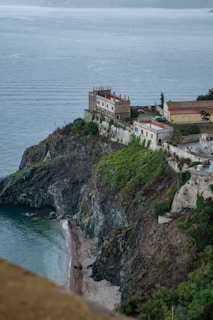 A coastal landscape featuring a rocky cliff with a series of buildings perched at the top. The structures have light-colored walls and red-tiled roofs, overlooking a narrow strip of beach with gentle waves. Lush greenery covers parts of the cliff, while the expansive sea stretches into the horizon.