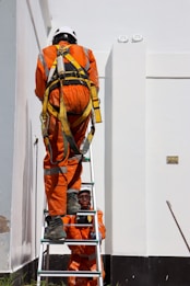 Two workers in bright orange safety suits and helmets are using a ladder against a large white building. One worker is climbing the ladder while the other steadies it from below. The workers are equipped with safety harnesses.