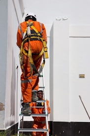 Two workers in bright orange safety suits and helmets are using a ladder against a large white building. One worker is climbing the ladder while the other steadies it from below. The workers are equipped with safety harnesses.