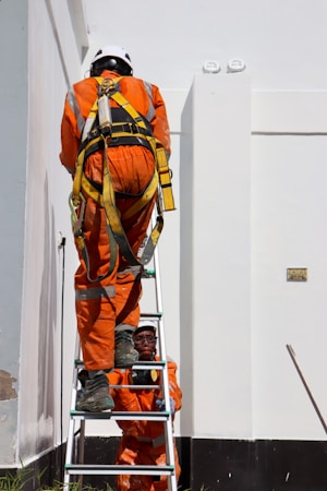 Two workers in bright orange safety suits and helmets are using a ladder against a large white building. One worker is climbing the ladder while the other steadies it from below. The workers are equipped with safety harnesses.