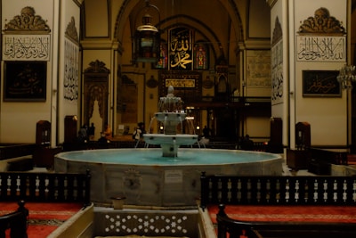 A large, ornate indoor space featuring Islamic architecture. In the center is a fountain with multiple tiers, surrounded by decorative railings. The walls are adorned with intricate Arabic calligraphy and archways, while hanging lamps provide a warm, ambient light. Red carpeted areas are visible, with some people scattered around the edges, suggesting a place of gathering or worship.