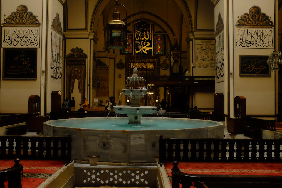 The image depicts a serene and ornate interior space, likely a mosque or a similar religious establishment. The focal point is a large fountain situated in the center of the room, surrounded by several benches arranged around it. The walls are adorned with intricate carvings and calligraphy, adding to the overall grandeur of the space. A chandelier hangs from the ceiling, casting a warm glow throughout the room. The floor is covered in tiles that reflect the light, enhancing the ambiance of the space.