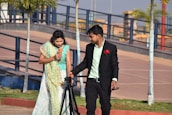 A man in a formal suit is holding a camera tripod and standing next to a woman in traditional attire, who is holding a flower. They appear to be in an outdoor park setting with trees and a paved walkway.