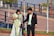 A man in a formal suit is holding a camera tripod and standing next to a woman in traditional attire, who is holding a flower. They appear to be in an outdoor park setting with trees and a paved walkway.