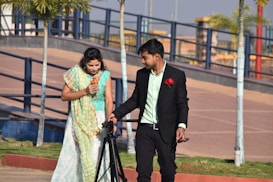 A man in a formal suit is holding a camera tripod and standing next to a woman in traditional attire, who is holding a flower. They appear to be in an outdoor park setting with trees and a paved walkway.