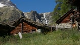 Charming rustic wooden cabins are nestled in a lush green meadow with a backdrop of towering rocky mountains. A narrow, cascading waterfall can be seen in the distance under a clear blue sky.