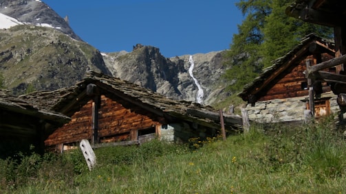 Charming rustic wooden cabins are nestled in a lush green meadow with a backdrop of towering rocky mountains. A narrow, cascading waterfall can be seen in the distance under a clear blue sky.