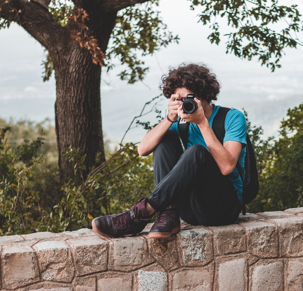 woman in blue shirt and black pants sitting on concrete wall holding camera during daytime