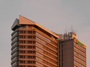 A tall building with a modern architectural design, featuring multiple floors with light-colored paneling and large windows. There is a sign for a Holiday Inn at the top of the structure, and various antennas and equipment are visible on the roof. The building is bathed in the warm hues of a setting or rising sun, casting an orange glow.