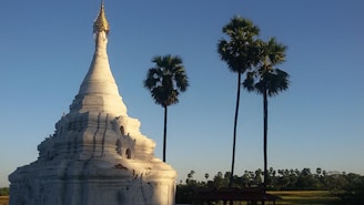 A white stupa with a gilded top stands prominently against a clear blue sky. Tall palm trees are positioned next to the stupa, casting long shadows. The background features distant trees and a flat landscape bathed in sunlight.