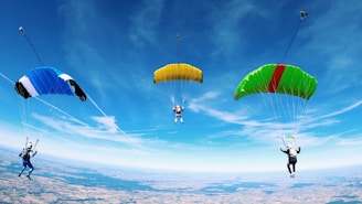Three skydivers are mid-air, each with a brightly colored parachute against a clear blue sky. The parachutes are blue, yellow, and green with a red stripe. The landscape below is visible, consisting of patches of land resembling fields or countryside.