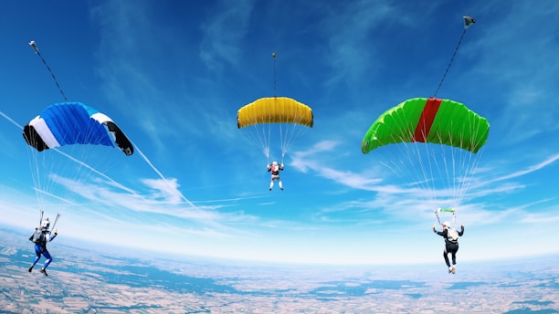 Three skydivers are mid-air, each with a brightly colored parachute against a clear blue sky. The parachutes are blue, yellow, and green with a red stripe. The landscape below is visible, consisting of patches of land resembling fields or countryside.