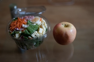A fresh salad in a clear plastic container, featuring ingredients like shredded carrots, lettuce, cheese cubes, sliced egg, and deli meat. Next to it is an apple, light red with a hint of yellow, resting on a flat surface.