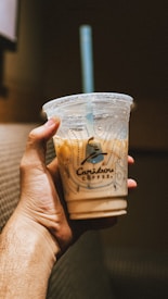 A hand holding a clear plastic cup with a dome lid filled with iced coffee, with a blue straw. The cup is branded with Caribou Coffee logo, featuring a stylized caribou. The background is softly lit and blurred, adding a cozy ambiance.