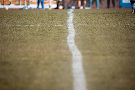 Close-up of a football on the grass field with a blurred scoreboard in the background.