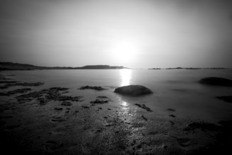 A black and white landscape photograph capturing a serene seascape during sunset or sunrise. The horizon is visible with distant landmasses, and the foreground features a rocky shoreline with smooth water reflecting the light.