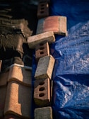 A set of bricks and roofing tiles are arranged on a surface that is divided into two parts. One side is covered with a blue tarp, and the other side is black. The bricks are of varying colors, mostly red and brown, and include some with holes.
