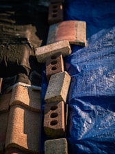 A set of bricks and roofing tiles are arranged on a surface that is divided into two parts. One side is covered with a blue tarp, and the other side is black. The bricks are of varying colors, mostly red and brown, and include some with holes.