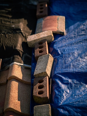 A set of bricks and roofing tiles are arranged on a surface that is divided into two parts. One side is covered with a blue tarp, and the other side is black. The bricks are of varying colors, mostly red and brown, and include some with holes.