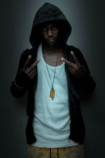 A person with a serious expression wearing a dark hoodie over a white tank top. They are making hand signs with both hands, displaying a peace sign. A necklace with a pendant hangs around their neck. The background is a plain dark color.