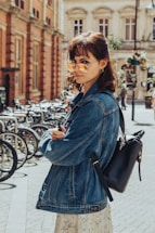 A person wearing a denim jacket and sunglasses stands on a city street lined with bicycles. The sun casts a warm glow, highlighting their stylish outfit and the cobblestone path beneath. The backdrop includes historic buildings and a row of parked bicycles, suggesting an urban setting.