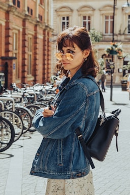 A person wearing a denim jacket and sunglasses stands on a city street lined with bicycles. The sun casts a warm glow, highlighting their stylish outfit and the cobblestone path beneath. The backdrop includes historic buildings and a row of parked bicycles, suggesting an urban setting.
