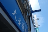 A blue outdoor sign with white text, featuring Asian characters and the word 'PHARMACY'. It's mounted on a building facade, surrounded by other signs and what appears to be an air conditioning unit.
