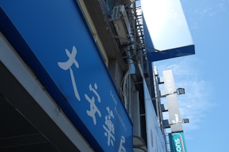 A blue outdoor sign with white text, featuring Asian characters and the word 'PHARMACY'. It's mounted on a building facade, surrounded by other signs and what appears to be an air conditioning unit.