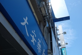 A blue outdoor sign with white text, featuring Asian characters and the word 'PHARMACY'. It's mounted on a building facade, surrounded by other signs and what appears to be an air conditioning unit.