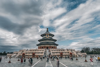 A large, ornate temple structure rises in the center of the image, characterized by its circular design and layered roofing. The temple is surrounded by a wide, open paved area with numerous people walking, taking photos, and exploring the surroundings. The sky above is filled with dramatic clouds, hinting at possible rain or clearing weather. The structure has traditional architectural details with intricate designs, and the atmosphere is busy yet calm.