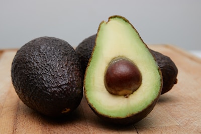 Two whole avocados and a halved avocado are placed on a wooden surface. The halved avocado displays its creamy green flesh and large brown seed.