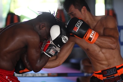 Two boxers are engaged in an intense boxing match inside a gym. The fighter on the left is wearing red shorts and defending with black and white gloves, while the fighter on the right is delivering a punch, wearing black and orange gloves with matching shorts. The background features boxing equipment and blurred shapes, indicating a training environment.