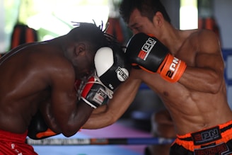 Two boxers are engaged in an intense boxing match inside a gym. The fighter on the left is wearing red shorts and defending with black and white gloves, while the fighter on the right is delivering a punch, wearing black and orange gloves with matching shorts. The background features boxing equipment and blurred shapes, indicating a training environment.