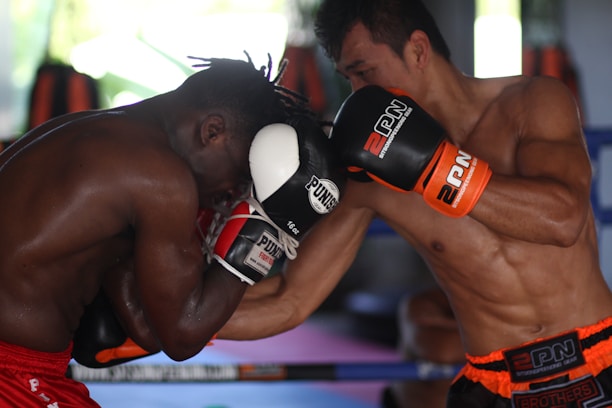 Two boxers are engaged in an intense boxing match inside a gym. The fighter on the left is wearing red shorts and defending with black and white gloves, while the fighter on the right is delivering a punch, wearing black and orange gloves with matching shorts. The background features boxing equipment and blurred shapes, indicating a training environment.