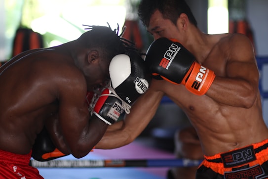 Two boxers are engaged in an intense boxing match inside a gym. The fighter on the left is wearing red shorts and defending with black and white gloves, while the fighter on the right is delivering a punch, wearing black and orange gloves with matching shorts. The background features boxing equipment and blurred shapes, indicating a training environment.