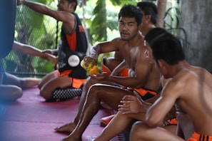 A group of men are sitting on a mat in a gym setting, wearing athletic shorts, conversing and possibly taking a break. One man holds an orange beverage bottle, indicating hydration during or after exercise. The background includes a fenced area with greenery visible through it.
