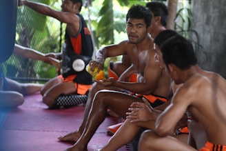 A group of men are sitting on a mat in a gym setting, wearing athletic shorts, conversing and possibly taking a break. One man holds an orange beverage bottle, indicating hydration during or after exercise. The background includes a fenced area with greenery visible through it.