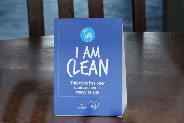 A blue table sign displays the text 'I AM CLEAN' with additional smaller text stating 'This table has been sanitised and is ready to use.' The sign includes a circular logo at the top depicting a hand with bubbles.